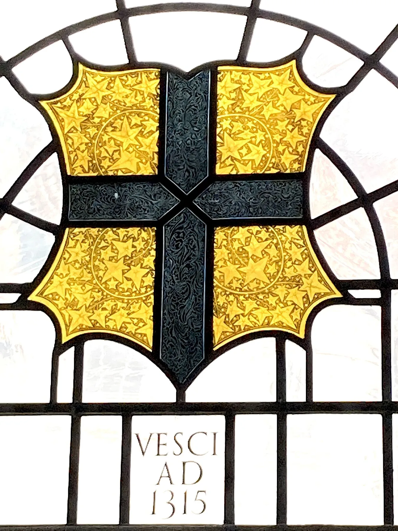A gold heraldic shield with a black cross, set in plain glass